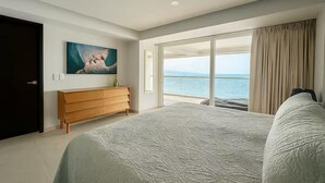 Quarto com uma cama grande, penteadeira de madeira, um quadro e vista para o mar através de uma porta de vidro deslizante.