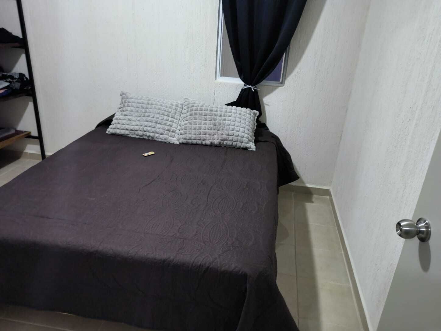 A small, neatly made bed with a dark bedspread and two pillows, a small shelf with folded clothes, and a window with a black curtain.