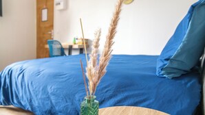 A room with a bed, a blue bedspread, a wooden headboard, a blue pillow, a green vase with dried plants, and a blue chair. - Charming, light-filled studio - Centre-ville-Gare (Cambrai)