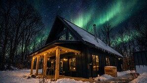 A wooden cabin with a covered porch, surrounded by snow, under a starry night sky with the aurora borealis.