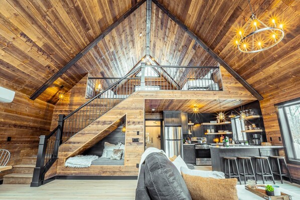 A spacious loft-style living area with a high ceiling, wooden walls, a staircase, a kitchen, and a bar area.