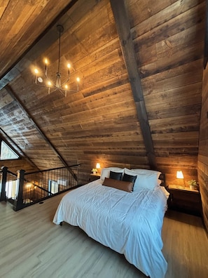 A spacious attic bedroom with a wooden ceiling, a large bed with white bedding, two bedside lamps, and a chandelier.