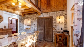 Un interior rústic amb una paret de pedra, bigues de fusta al sostre, una taula de dinar de fusta amb cadirees i una llar de pedra.