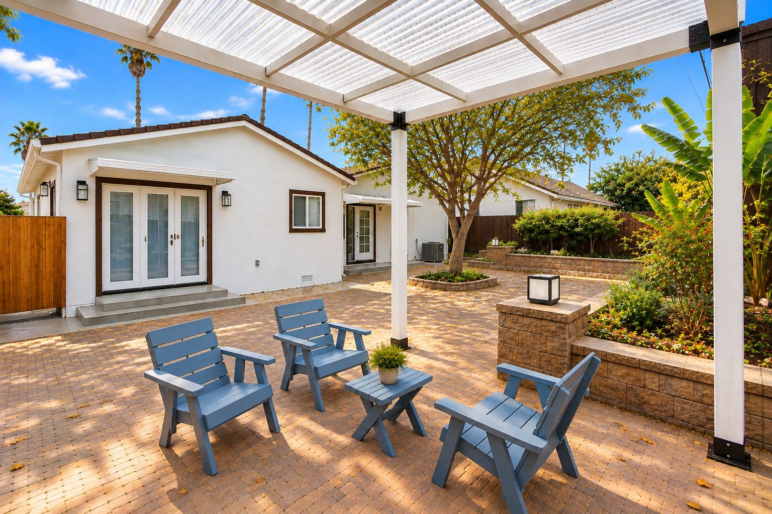 A covered patio area with blue chairs and a table, a white house with a garage, and a garden with various plants.