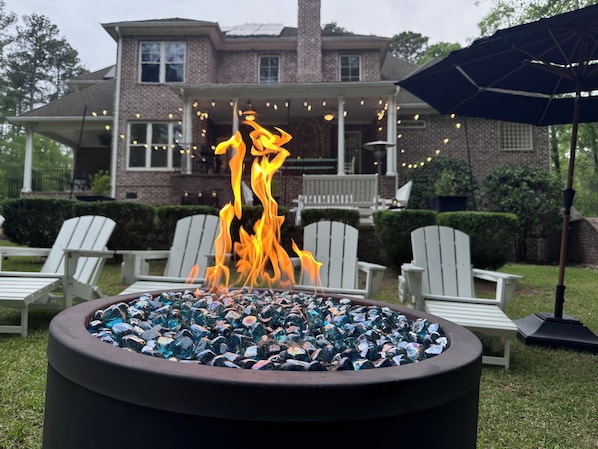 A fire pit with stones, surrounded by white Adirondack chairs, in front of a brick house with a porch and string lights.