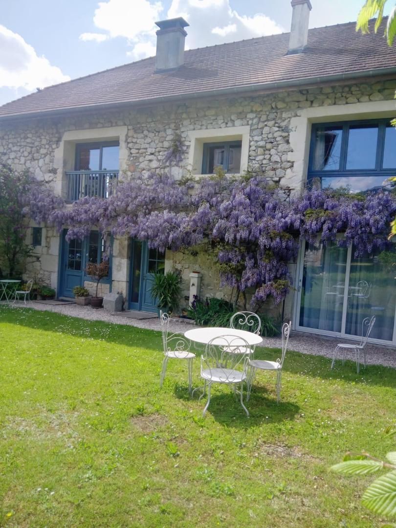 Une maison en pierre dotée d’un balcon, de plantes grimpantes aux fleurs violettes et d’un petit espace extérieur aménagé avec des chaises en métal.