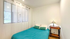 A bedroom with a bed, a nightstand, a lamp, and a window with curtains. - Une Maison Pour Deux (BILLIEME)