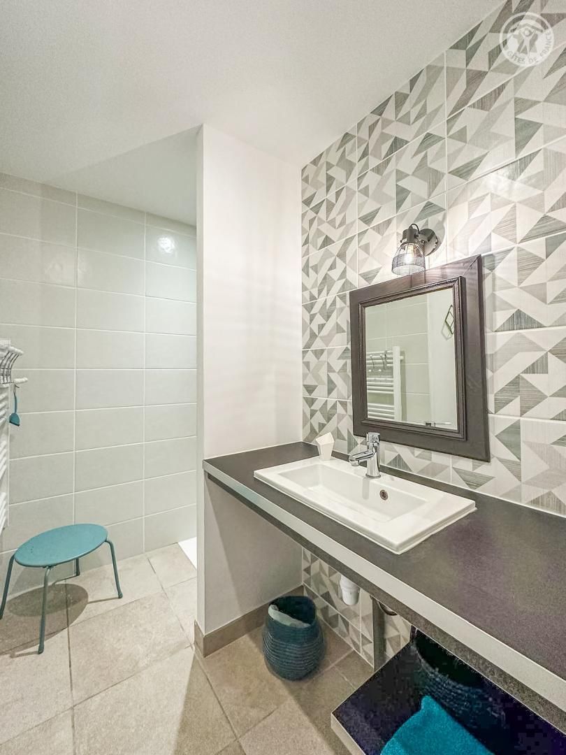 Baño con lavamanos blanco, un espejo y pared con diseño geométrico. Hay una silla azul y un cesto de basura.