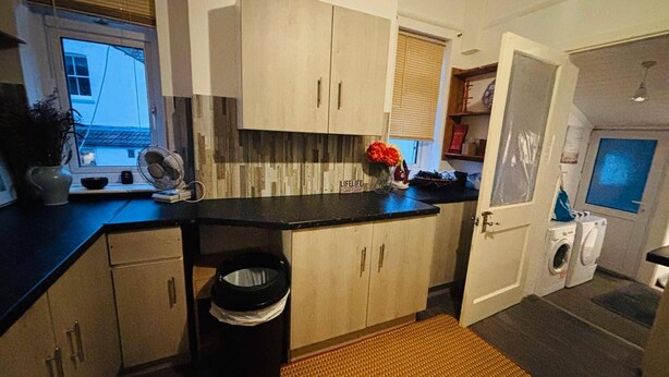 A compact kitchen with wooden cabinets, a black countertop, a small trash bin, and a window with blinds.