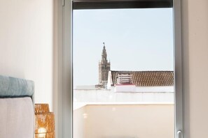 Vista per una finestra d'un edifici amb una torre destacada, teulada de rajola i una cadira de vimet.