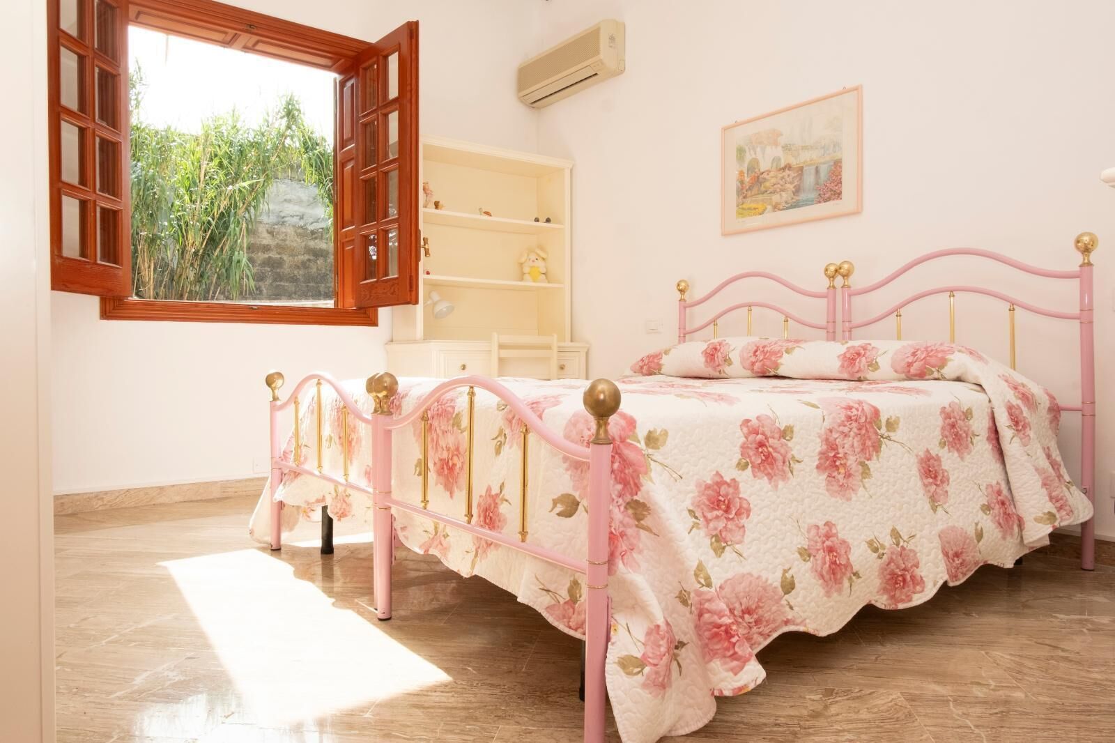 Een slaapkamer met een roze metalen bedframe, bloemenbeddengoed, een witte nachtkastje, een houten plank en een raam met een open luik.