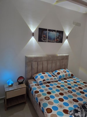 Une chambre à coucher avec un lit, une table de chevet, une lampe fixée au mur et un tableau encadré.