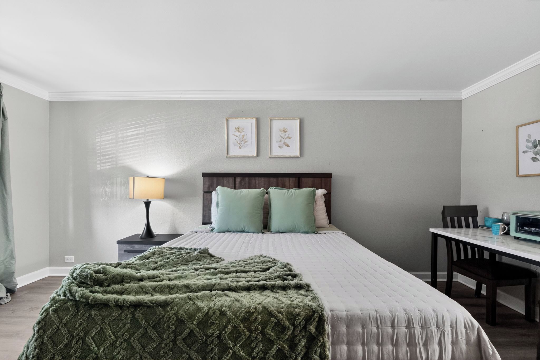 A bedroom with a bed, a nightstand with a lamp, a desk, and two framed pictures on the wall.