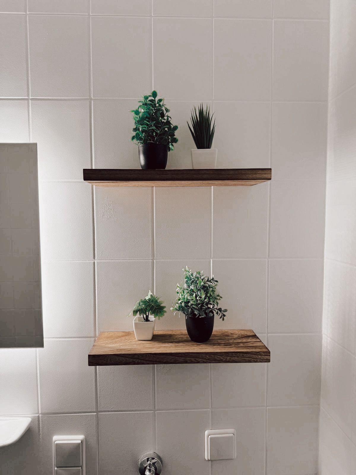 A bathroom with white tiled walls, two wooden shelves with potted plants, a white sink, and a toilet.
