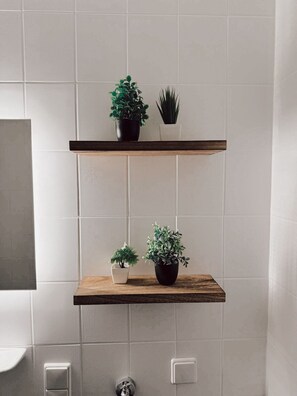 A bathroom with white tiled walls, two wooden shelves with potted plants, a white sink, and a toilet. - Vacation Apartment - Cozy Home by Li-Apartments (Pforzheim)