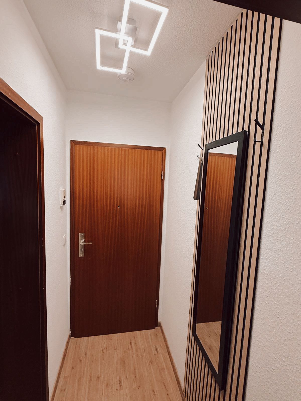 Un couloir avec une porte en bois, un miroir et un luminaire au plafond.