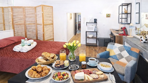 Une chambre d’hôtel comprenant un lit, une table pour le petit-déjeuner avec des croissants, des fruits, du fromage et du café, une kitchenette et une salle de bain avec douche et toilettes.