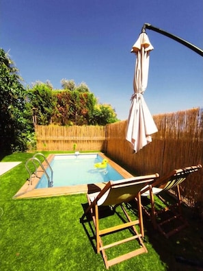 Un patio trasero con piscina, terraza de madera y una silla colgante.