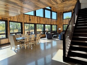 A spacious room with a wooden ceiling and large windows, a dining area with chairs and a table, and a staircase.