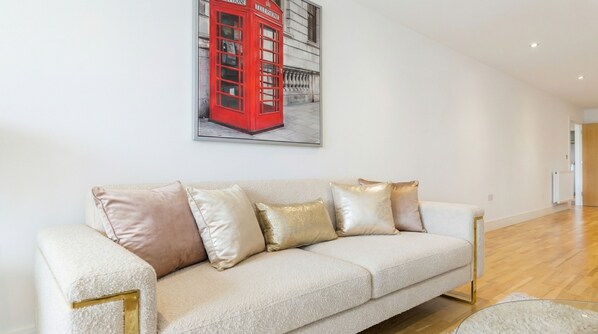 Een moderne woonkamer met een witte bank, gouden en beige kussens, en een lijstwerk met daarin een afbeelding van een rode telefooncel aan de muur.
