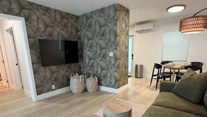 A modern living room with a stone-patterned wall, a flat-screen TV, a green sofa, wooden coffee tables, and a dining area with a table and chairs. - Renovated 1 Bedroom, Boutique Mid-Century Building (Fort Lauderdale)