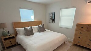 A bedroom with a large bed, bedside tables, a dresser, and two windows. - Renovated 1 Bedroom, Boutique Mid-Century Building (Fort Lauderdale)