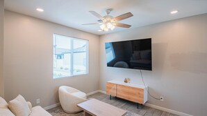 A modern living room with a ceiling fan, a flat-screen TV mounted on the wall, a light-colored sofa, a wooden coffee table, and a window with blinds. - The One 311 (Glendale)