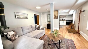 Ein modernes Wohnzimmer mit einem grauen Sofa, einem Couchtisch und einem Teppich. Im Hintergrund ist eine Küche mit weißen Schränken und einem hölzernen Barhocker zu sehen.