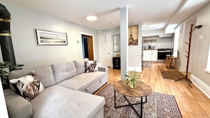 Ein modernes Wohnzimmer mit einem grauen Sofa, einem Couchtisch und einem Teppich. Im Hintergrund ist eine Küche mit weißen Schränken und einem hölzernen Barhocker zu sehen.