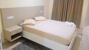 Ein ordentlich bezogenes Bett mit Kopfteil, ein Nachttisch mit Schublade und ein Vorhang am Fenster.