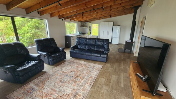 A spacious living area with a black leather sofa, a black leather chair, a large flat-screen TV, and a kitchen in the background.