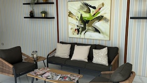 Un salon moderne avec un papier peint à rayures, une grande peinture abstraite, un canapé foncé, des fauteuils en osier et une table basse avec des magazines.