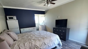 Een slaapkamer met een bed, een dressoir, een televisie, een plafondventilator, een magnetron en een raam.