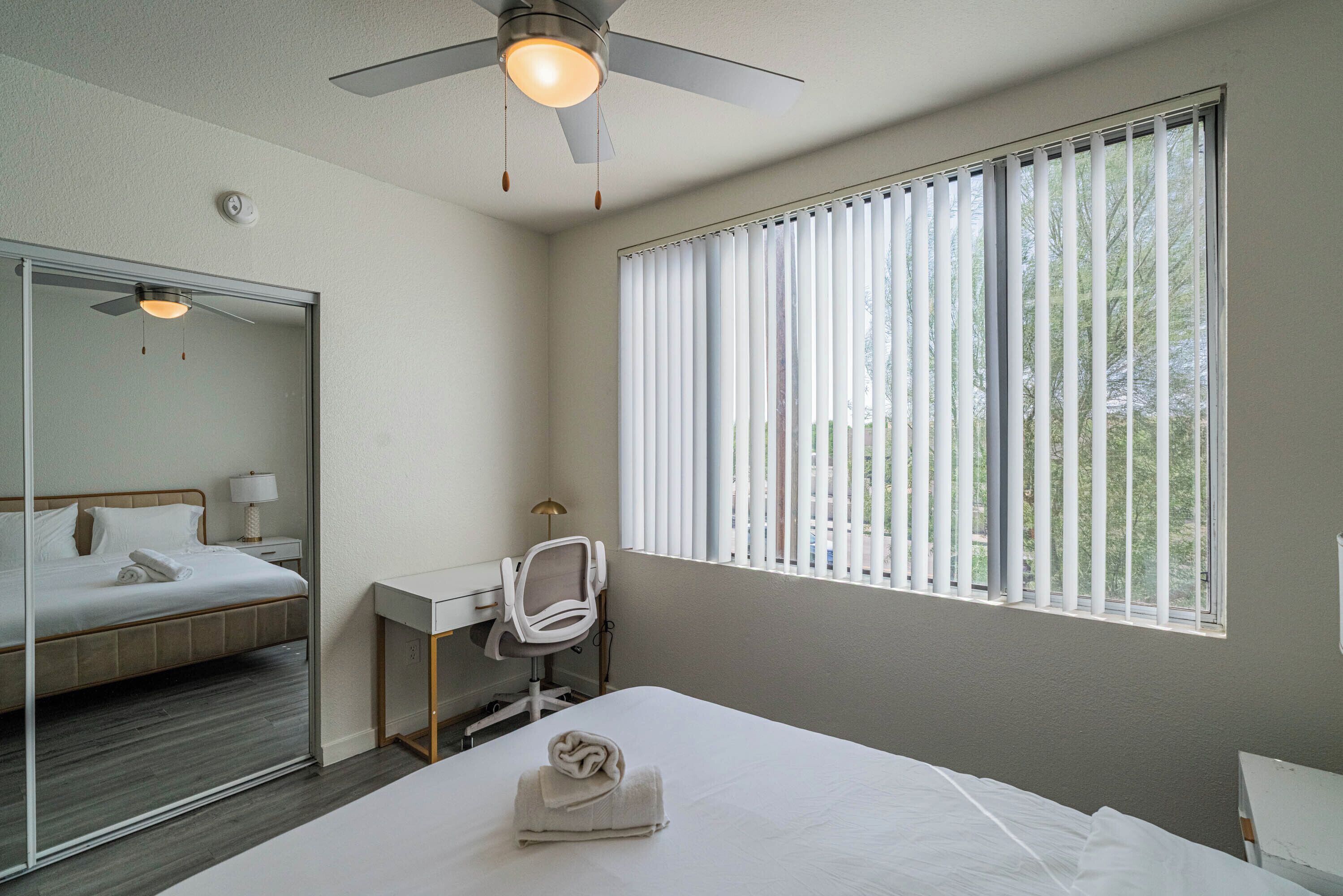 A hotel room with a bed, a desk, a chair, a ceiling fan, and a window with blinds.
