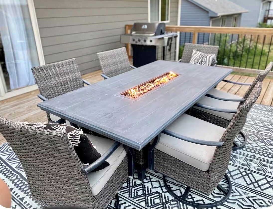 A rectangular outdoor table with a built-in fire feature, surrounded by wicker chairs with cushions.