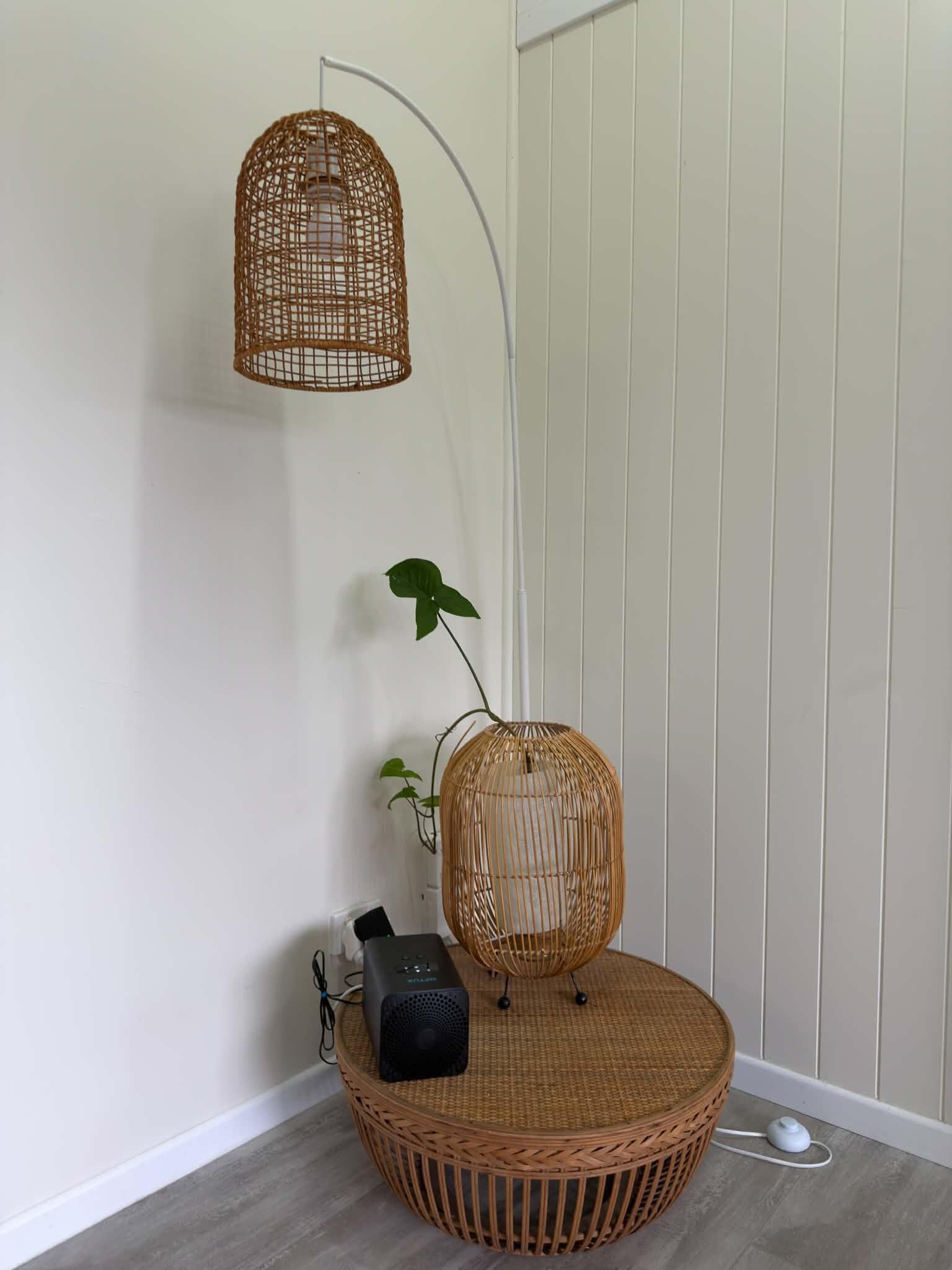 A room with a round wicker table, a black electronic device, a white wall, and a hanging wicker light fixture.