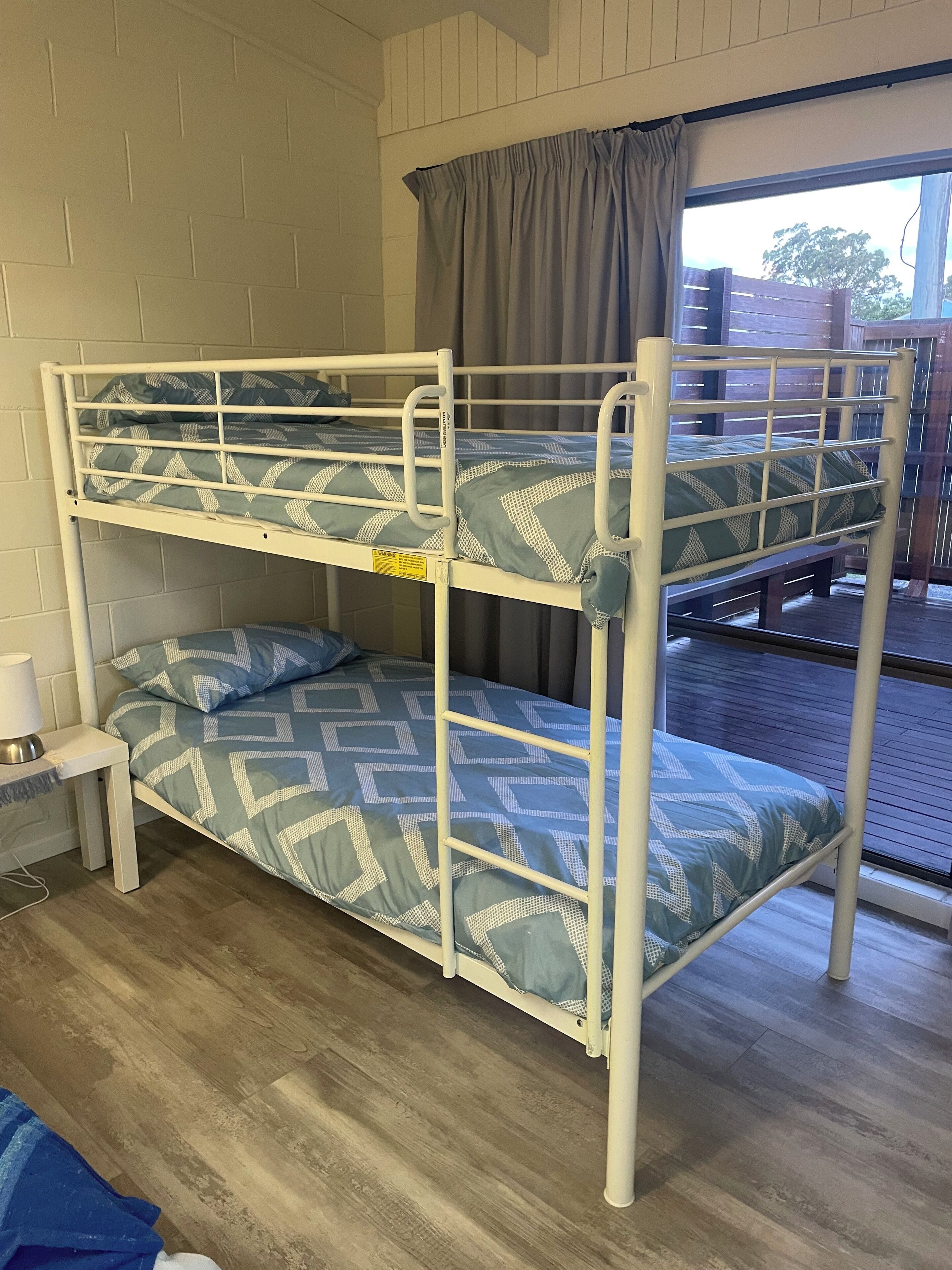 A bunk bed with a patterned blue and white bedding, a bedside table with a lamp, and a window with curtains.