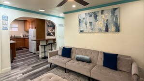 A spacious living room with a beige sofa, a matching ottoman, and a ceiling fan. A kitchen is visible in the background with wooden cabinets and a refrigerator. - Serene 2-bedroom condo in Santa Cruz near the beach (Santa Cruz)