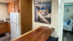 A compact kitchen with wooden cabinets, a white refrigerator, and a wooden dining table with black chairs. A large painting of a coastal scene is on the wall. - Serene 2-bedroom condo in Santa Cruz near the beach (Santa Cruz)