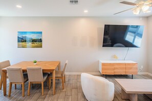 Un espace repas moderne avec une table et des chaises en bois, un téléviseur fixé au mur et un petit meuble orné d’un motif de feuilles.