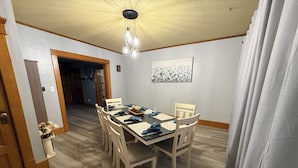 Une salle à manger avec une table en bois, des chaises et un lustre. On y trouve une porte qui mène à une autre pièce, ainsi qu’une fenêtre avec des rideaux.
