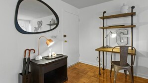 Una habitación con piso de madera, un escritorio con una lámpara, una silla, un estante decorado con una 'G' y un espejo en la pared.