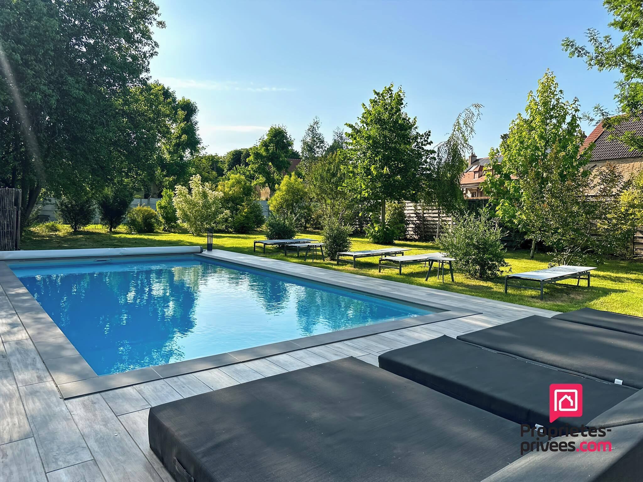 A rectangular swimming pool with a tiled deck, surrounded by green grass and trees. There are lounge chairs and a small table in the pool area.