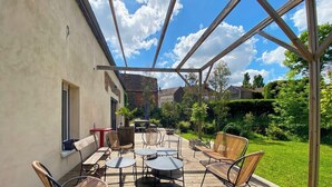 An outdoor patio with wooden furniture, a glass enclosure, and a view of a garden and buildings in the background. - Gîte with swimming pool at the gateway to the Morvan - Up to 15 people (cussy les forges)