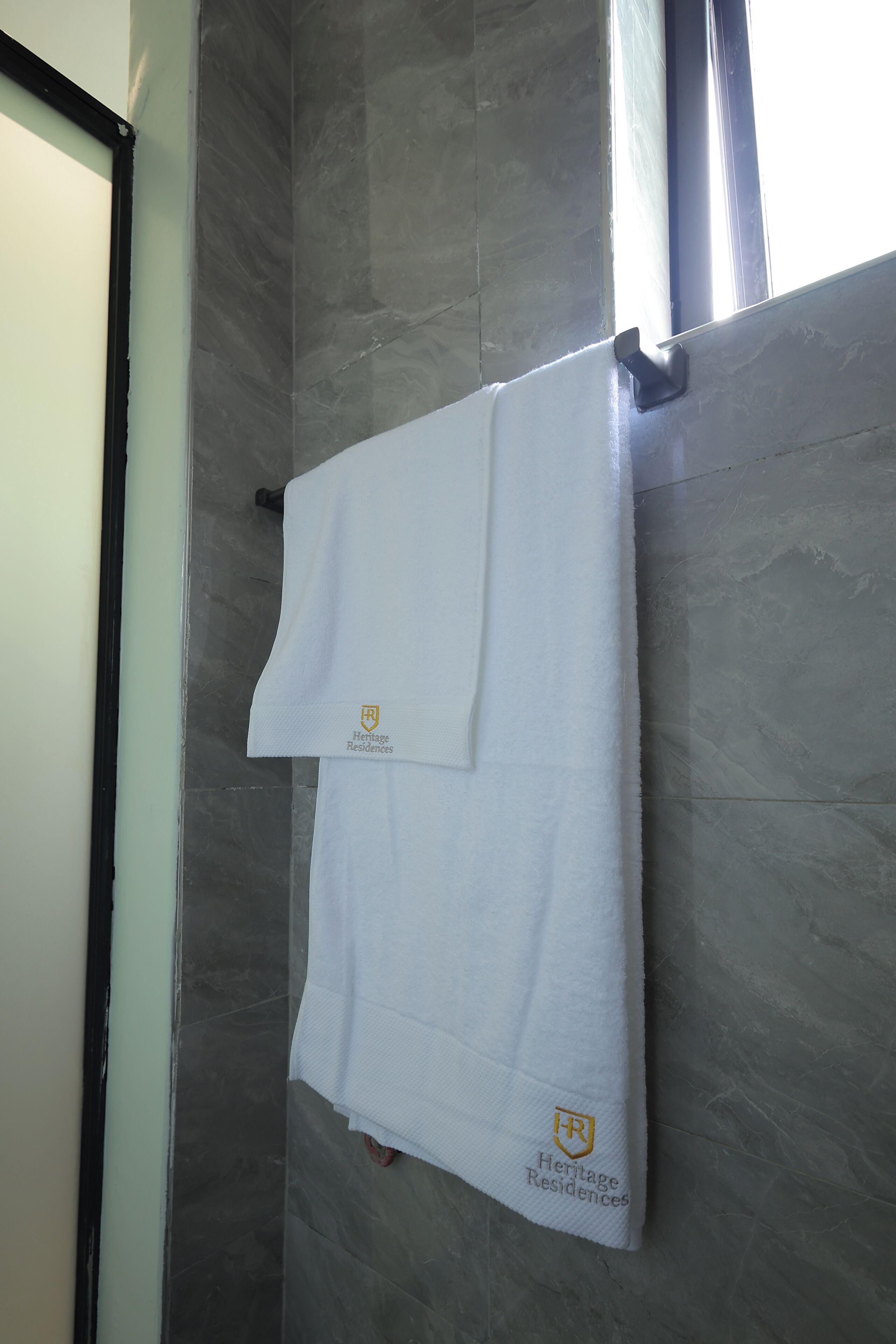 A white towel with "Heritage Residences" logo hanging on a metal rod in a bathroom with grey marble tiles.