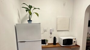 Een compacte keuken met een witte koelkast, een magnetron, een klein houten tafeltje en een potplant.