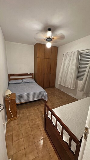 Ein kleines, gut beleuchtetes Schlafzimmer mit einem Einzelbett, einem Holzschrank, einem Deckenventilator und einem Fenster mit Vorhängen.