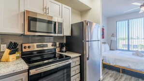 A modern kitchen with stainless steel appliances, a microwave, and a coffee maker. There is a bed with white bedding and a window with blinds. - Access 3011 (Tempe)