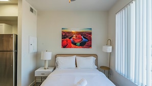 A hotel room with a bed, a nightstand, a lamp, a window with blinds, and a refrigerator. - Access 3011 (Tempe)