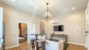 A spacious living area with a wooden floor, a central seating area with a table and chairs, a chandelier, a television mounted on the wall, and an air conditioning unit.